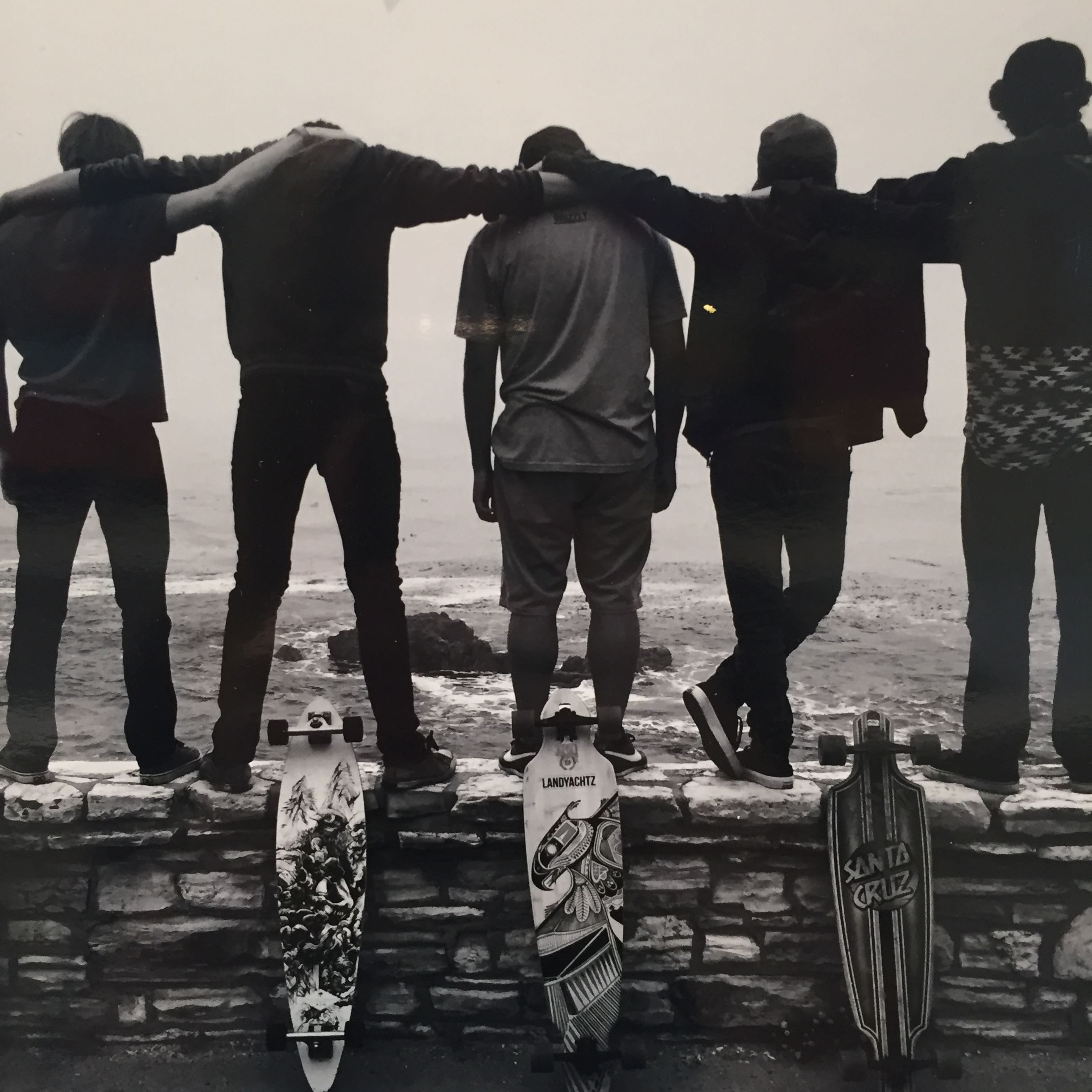 Black and white photo of three people with skateboards standing on brick wall facing the ocean